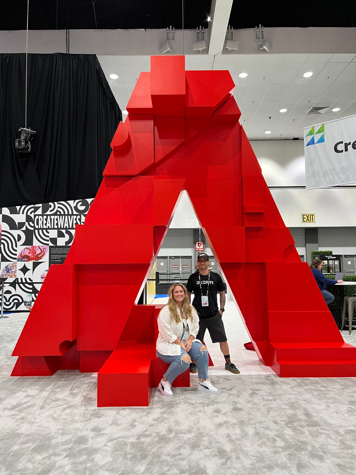 Two people posing in front of a large red Adobe logo sculpture at an Adobe event, with creative signage and booths in the background.