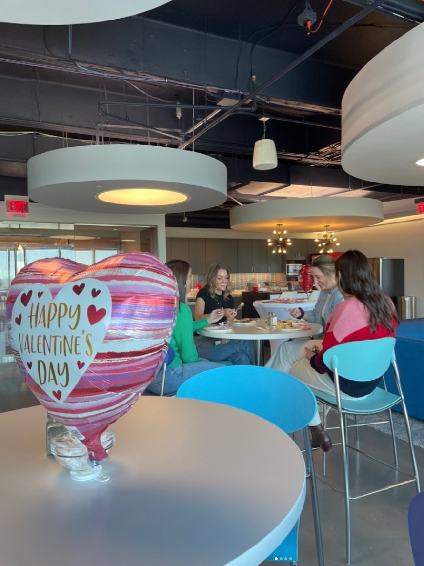 Four people eating at a round table in the Sagepath Reply office, with a 'Happy Valentine’s Day' heart balloon in the foreground.