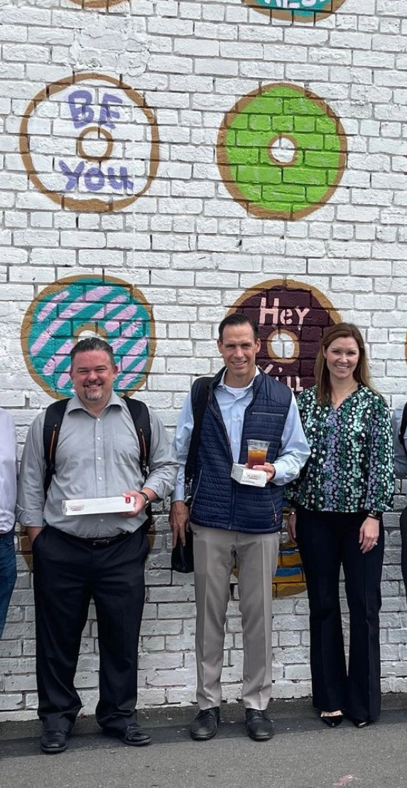 Three people smiling and holding Krispy Kreme donuts in front of a mural featuring colorful donuts with playful messages.