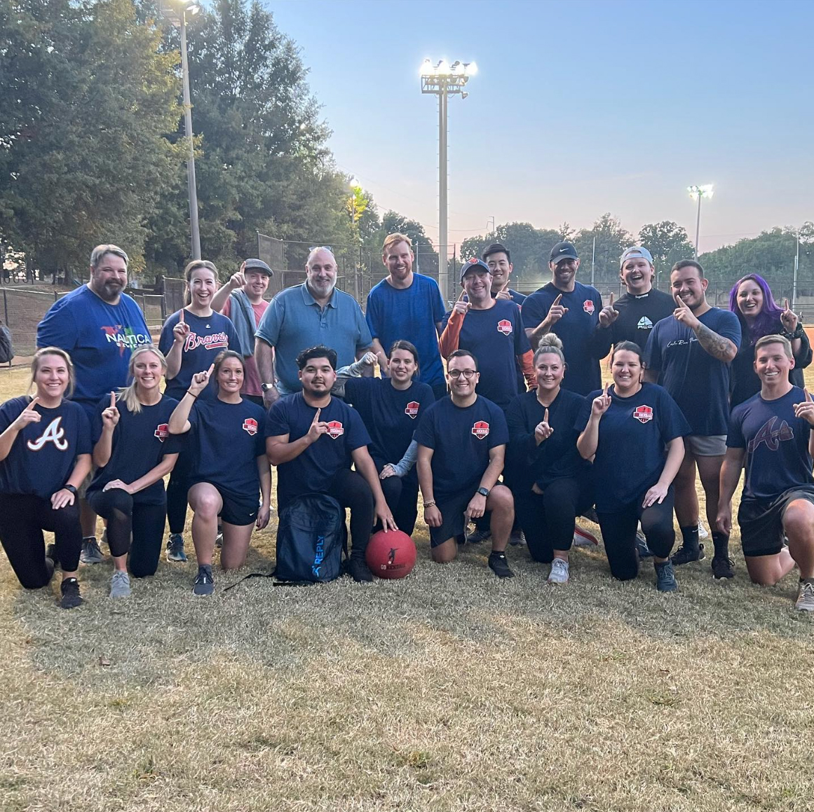 A group of employees posing together on a field as part of a company culture event, representing their kickball team, all smiling and holding up 'number one' hand signs.