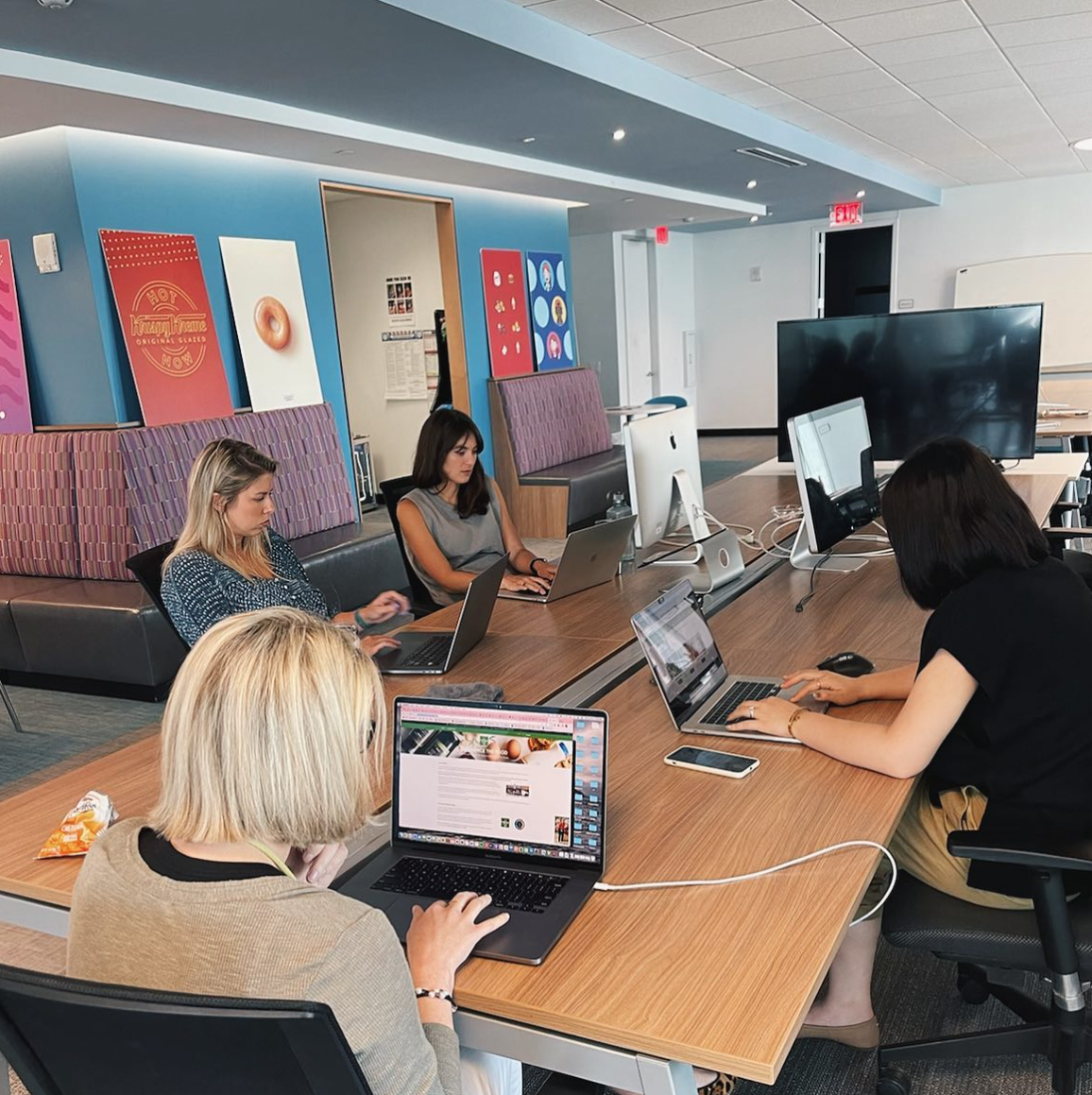 Four people working on laptops in the Sagepath Reply office, with vibrant donut-themed artwork on the walls and a collaborative workspace setup.