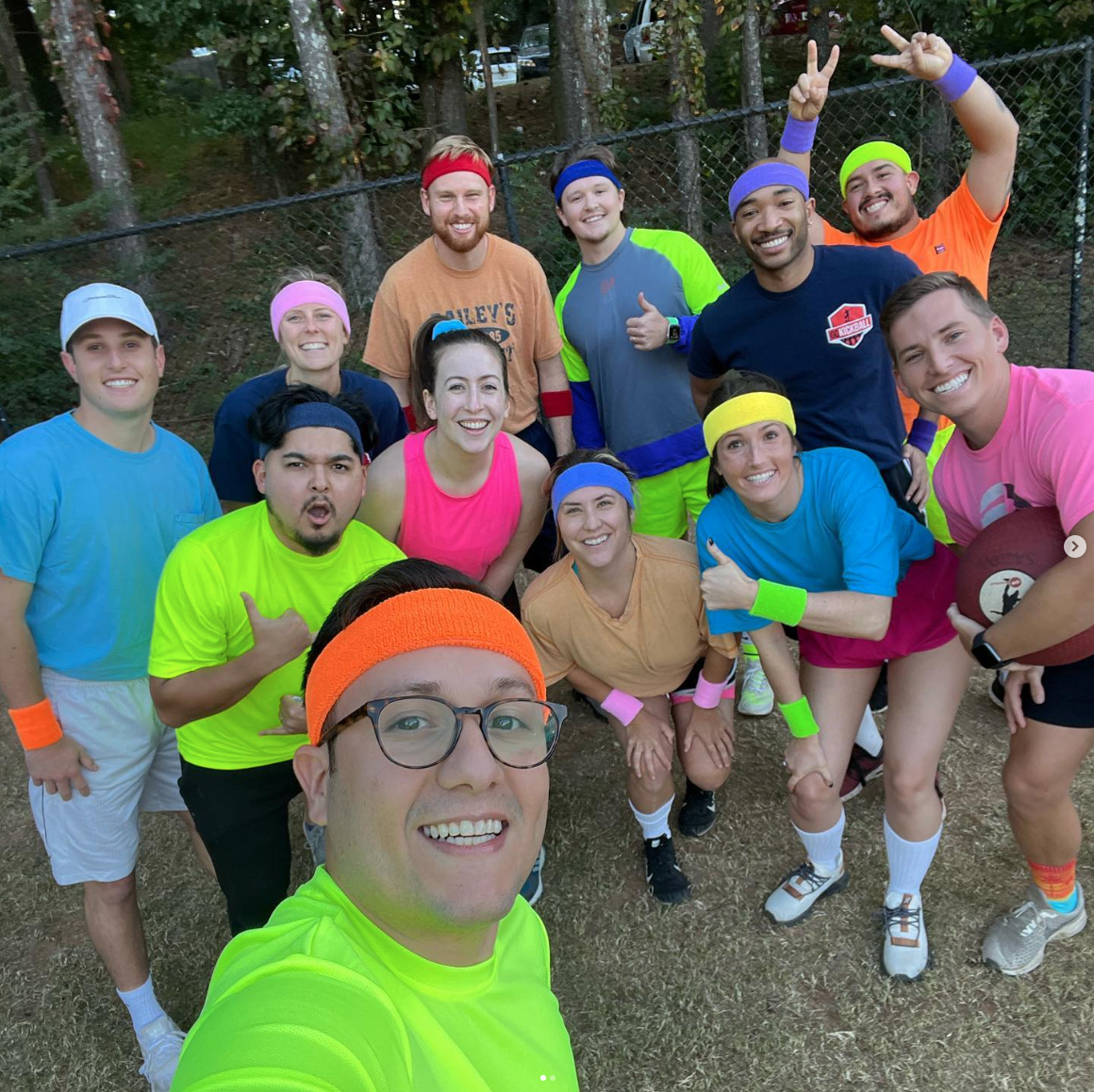 A group of employees in brightly colored athletic outfits posing together on a field, representing the Sagepath kickball team and highlighting a fun, vibrant company culture.