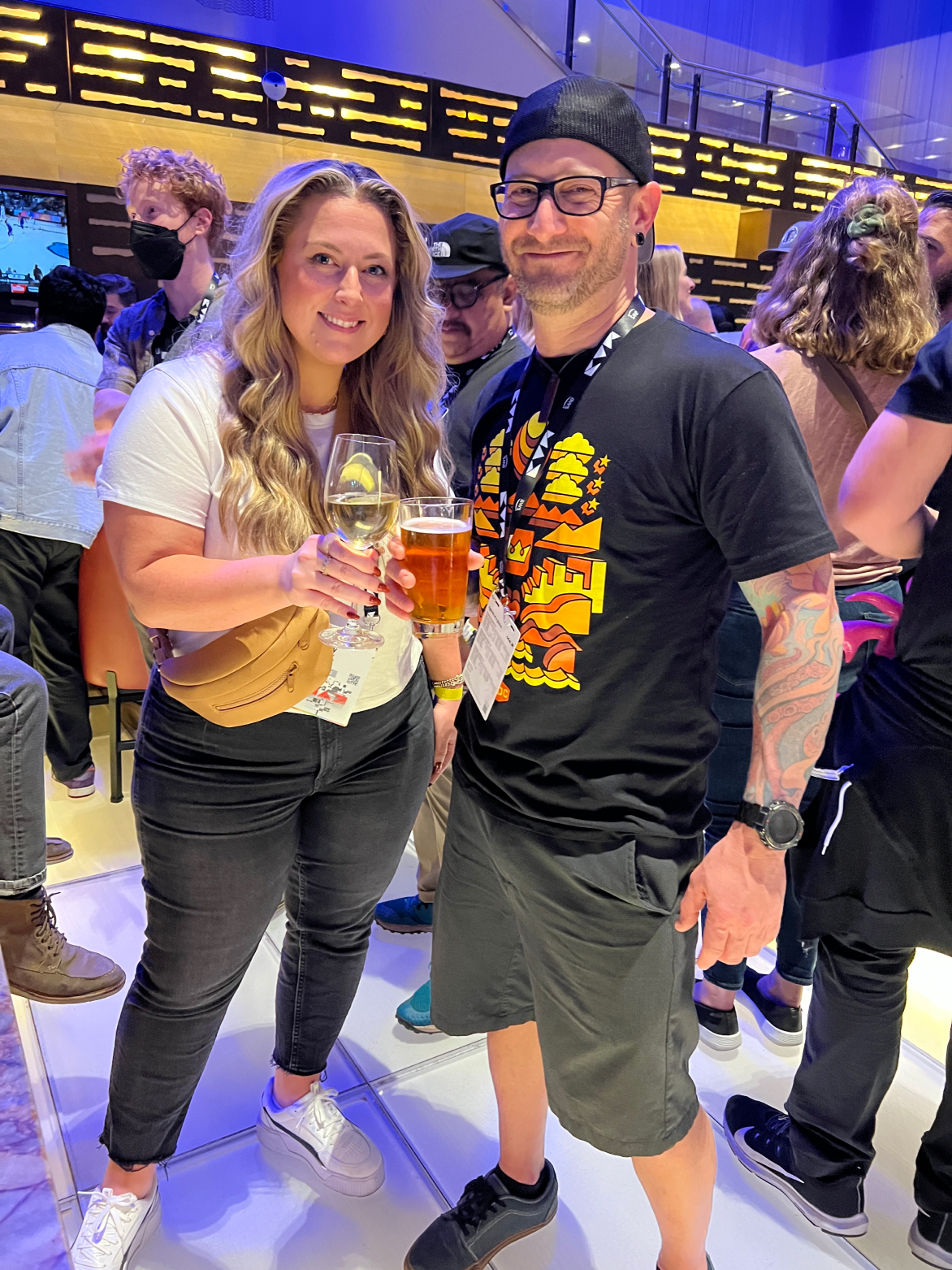 Two people smiling and holding drinks at a social event, with others in the background. One person holds a glass of wine, and the other holds a beer.