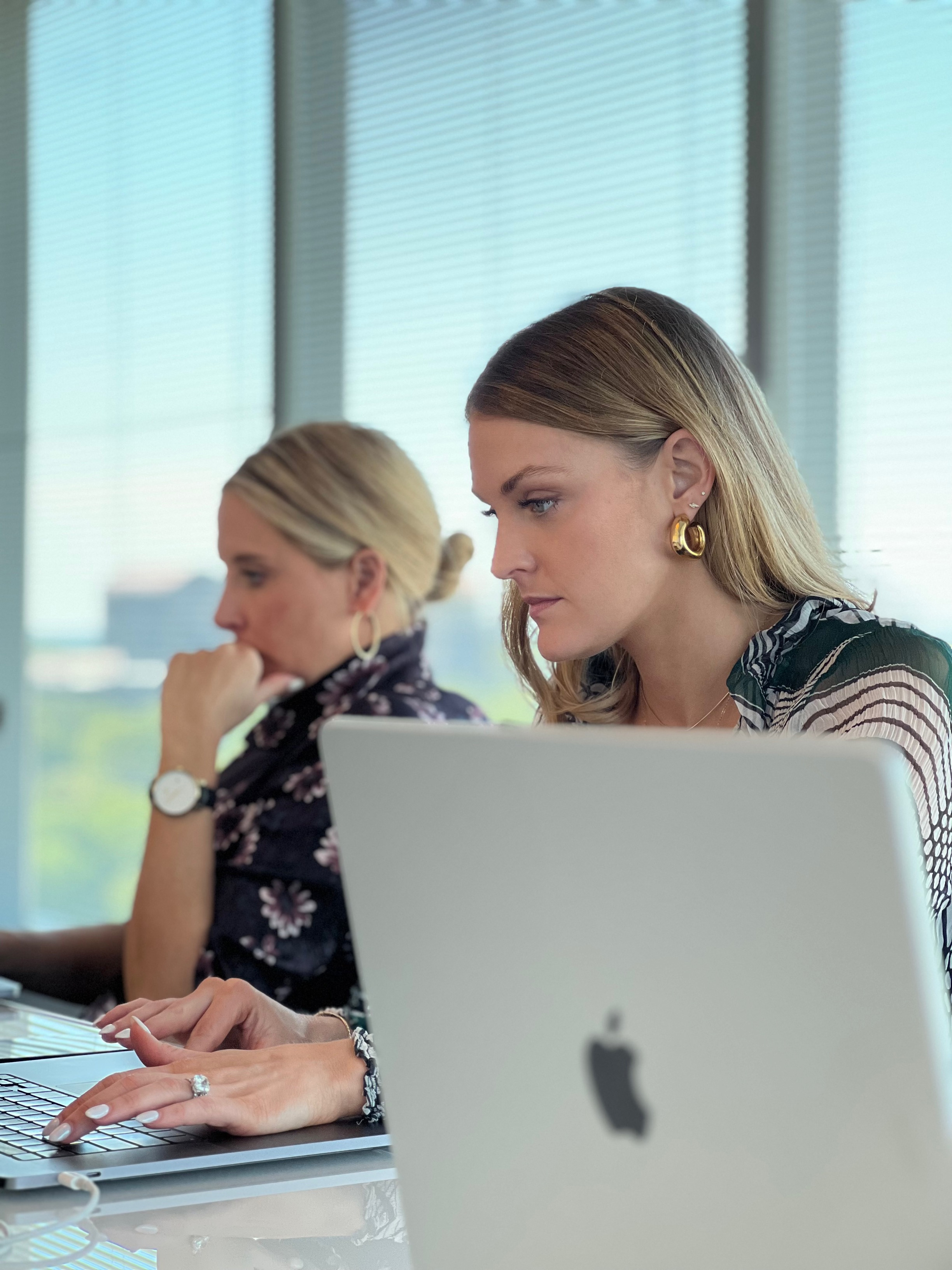 Two digital marketers working intently on laptops in a modern office with large windows, focused on their tasks.